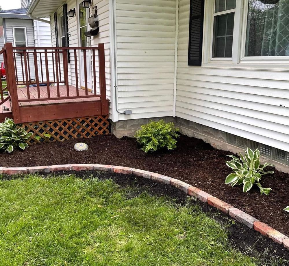 Landscape design featuring a curved brick border, fresh mulch, and vibrant greenery near a house.