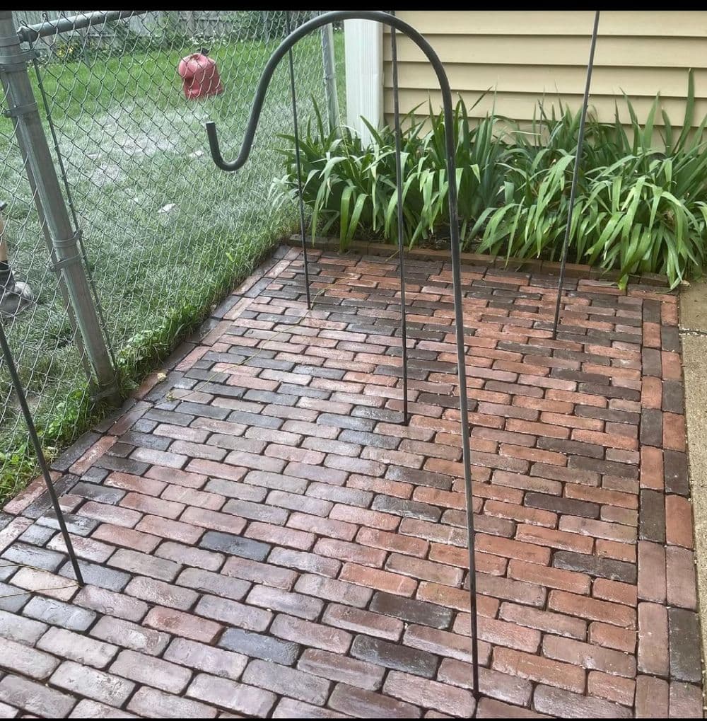 Metal garden hook on a brick patio surrounded by greenery and a fenced yard.