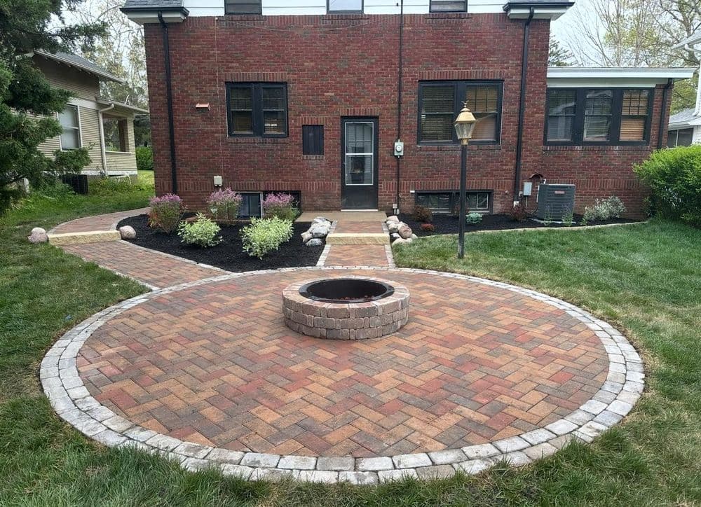 Brick patio with firepit and landscaped garden outside a brick house. Pathway and lamp included.