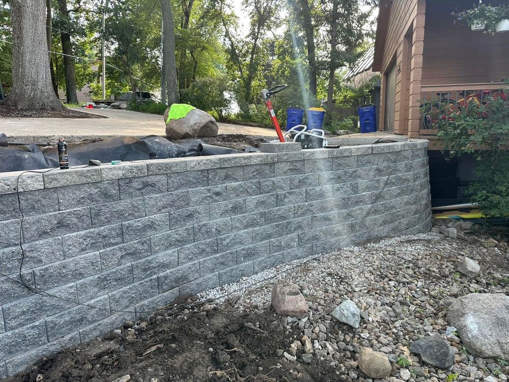 Newly built stone retaining wall in a landscaped yard with tools and rocks visible.