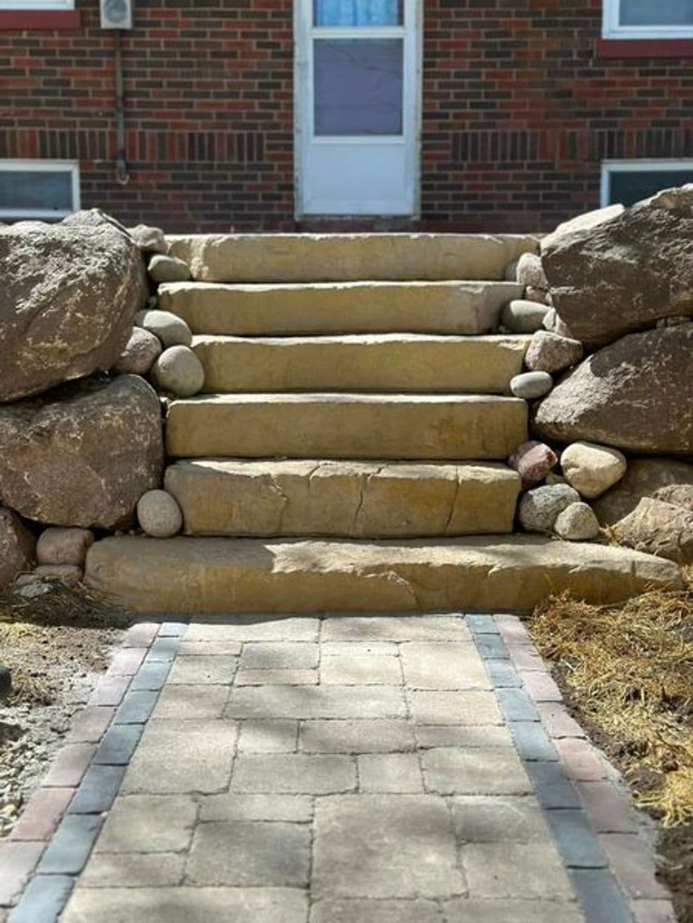 Stone steps framed by boulders leading to a front door, with a brick pathway in the foreground.