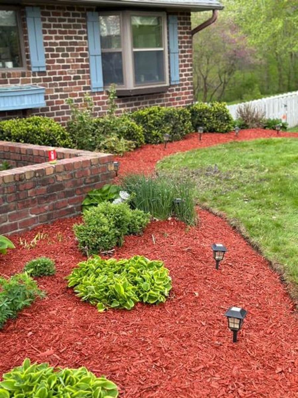 Vibrant landscaping with red mulch, lush greenery, and solar lights near a brick home.