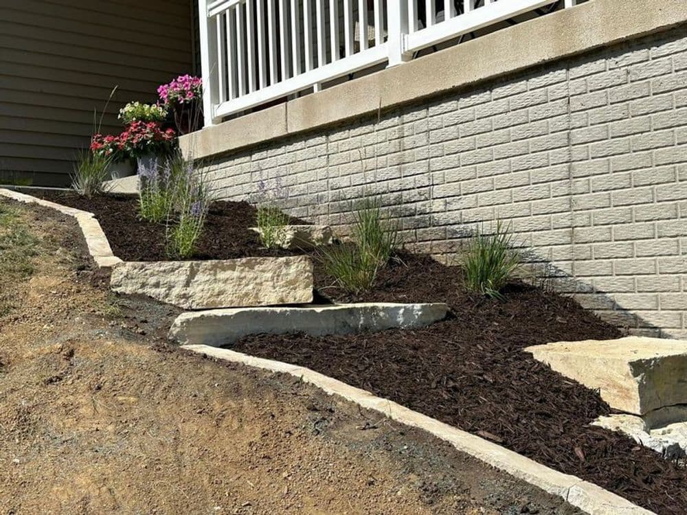 Landscaped garden with rocks, plants, and mulch along a house foundation.