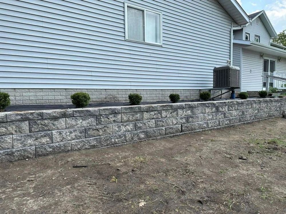 Stone retaining wall with neatly trimmed shrubs beside a light blue house.