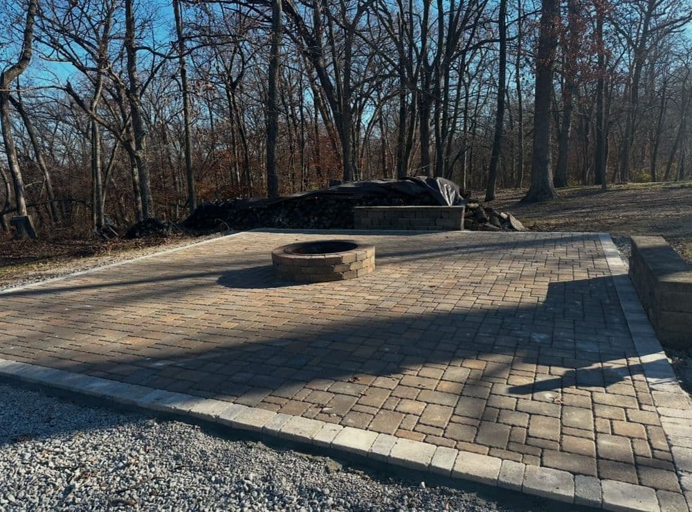 Paved outdoor fire pit area surrounded by trees and gravel in a wooded setting.