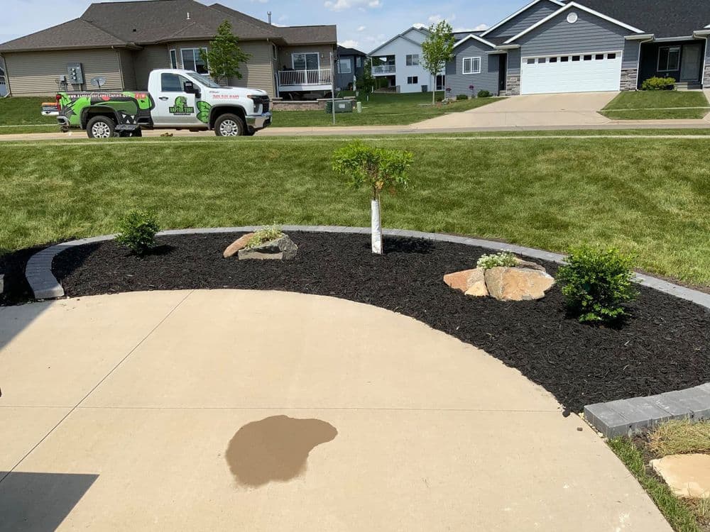 Landscaped yard with mulch, rocks, and small shrubs near a driveway and a service truck.