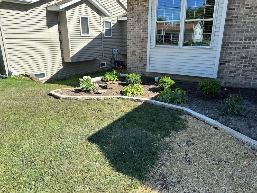 Lush garden bed featuring plants and stones beside a brick house in a sunny yard.
