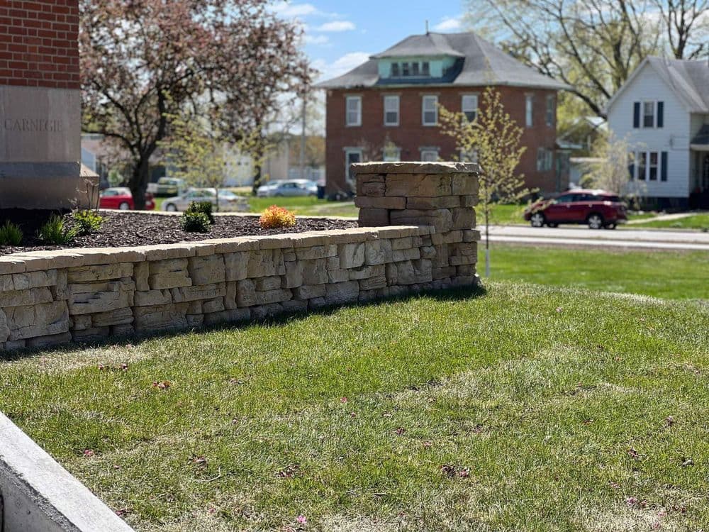 Stone landscape wall with flowers, green grass, and residential buildings in the background.